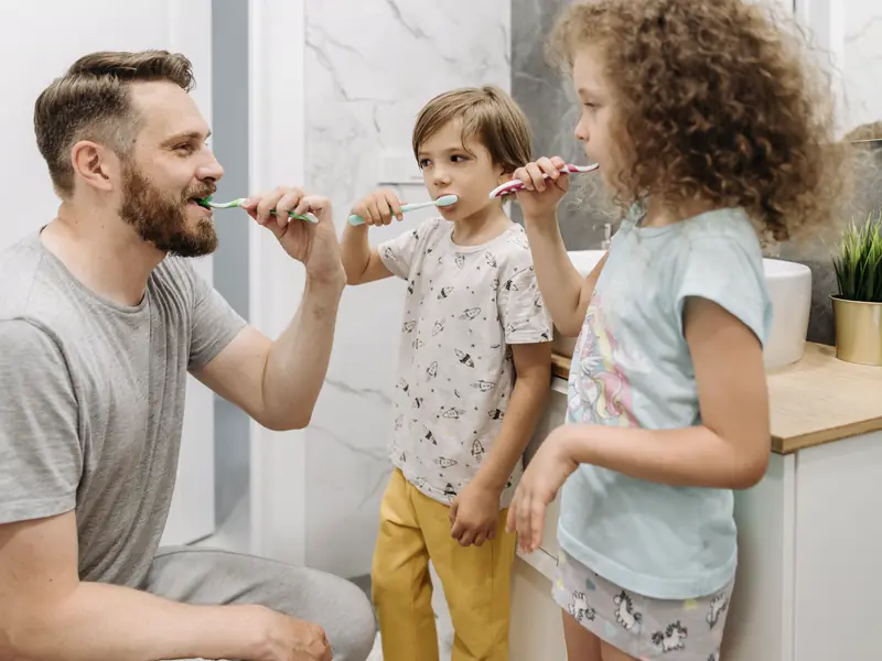 A dad and two kids brushing their teeth together in a bright bathroom