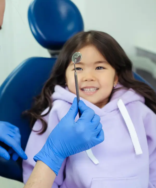 A smiling girl in a lilac hoodie sits in a blue dental chair while the dentist holds a small mirror