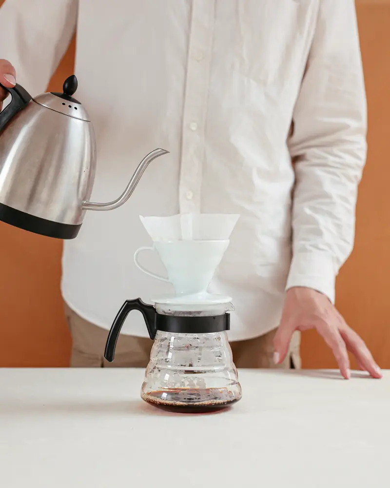A barista pours hot water from a gooseneck kettle into a white pour-over dripper