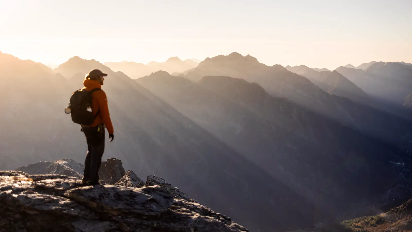 A hiker in an orange jacket stands on a rocky summit at sunrise, looking over ridges lit by sun rays