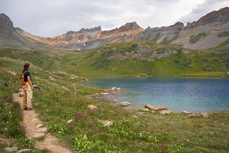 A hiker walks a dirt trail beside an alpine lake ringed by peaks