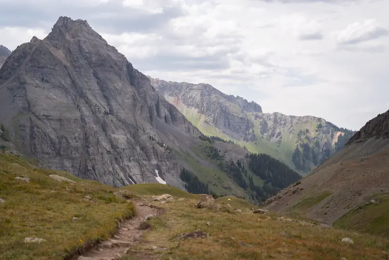 A narrow trail climbs a green valley toward a sharp gray peak