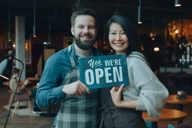Two cafe owners smiling and holding an open sign inside their shop