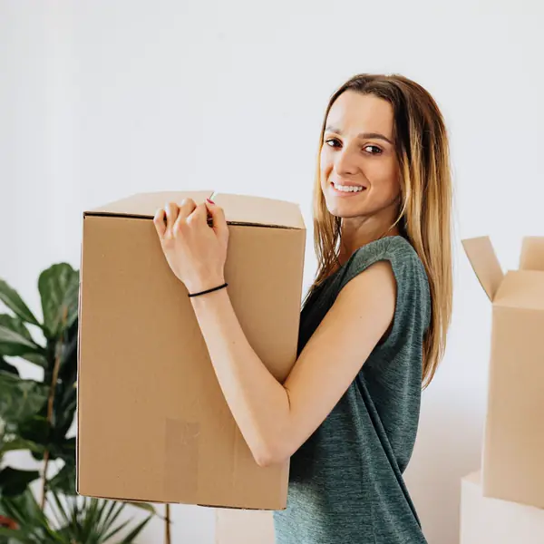 A smiling woman carries a moving box into her new apartment