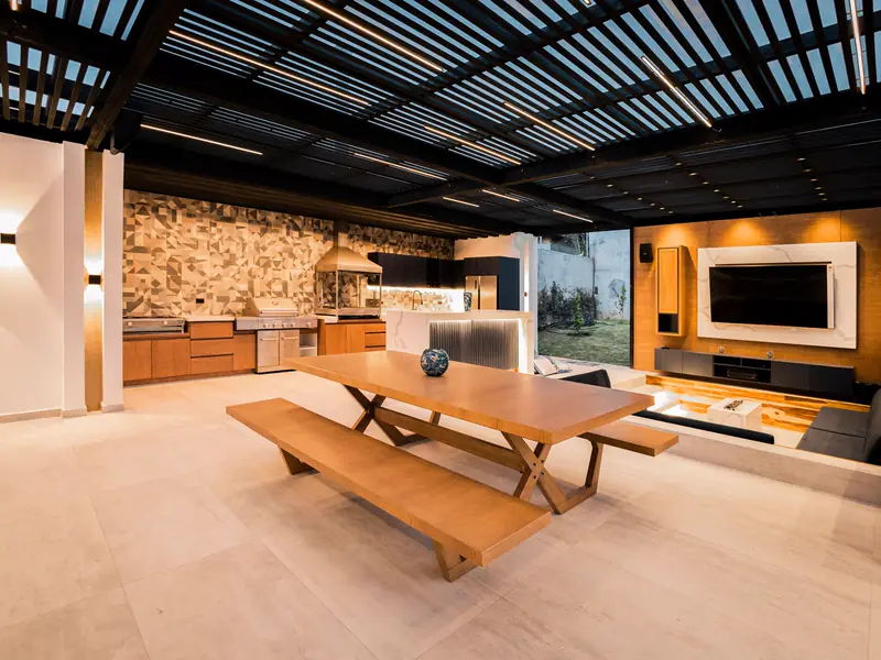 A covered outdoor kitchen and dining pavilion with a slatted roof and warm lighting