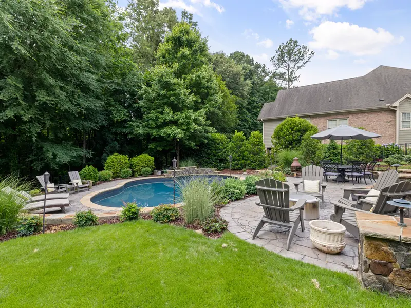 A pool with a stone surround, lounge chairs and dining table on a travertine terrace, framed by lawn and planting