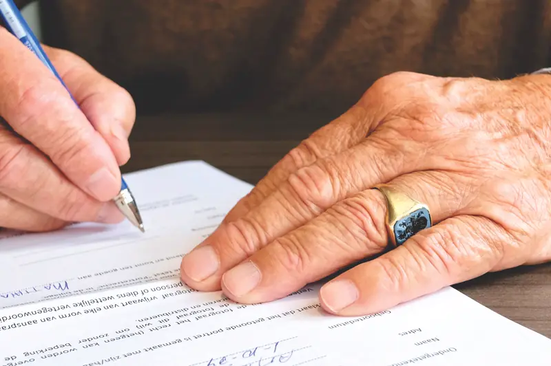An older man's hand rests on a document while another person signs it