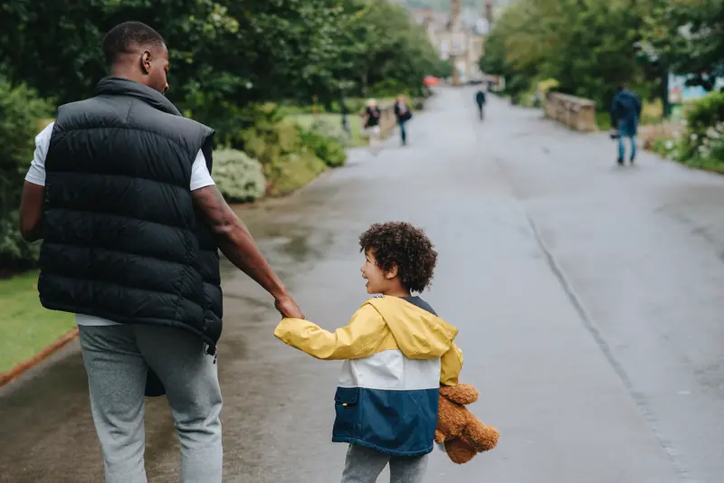 A father walks hand in hand with his young son down a tree-lined path