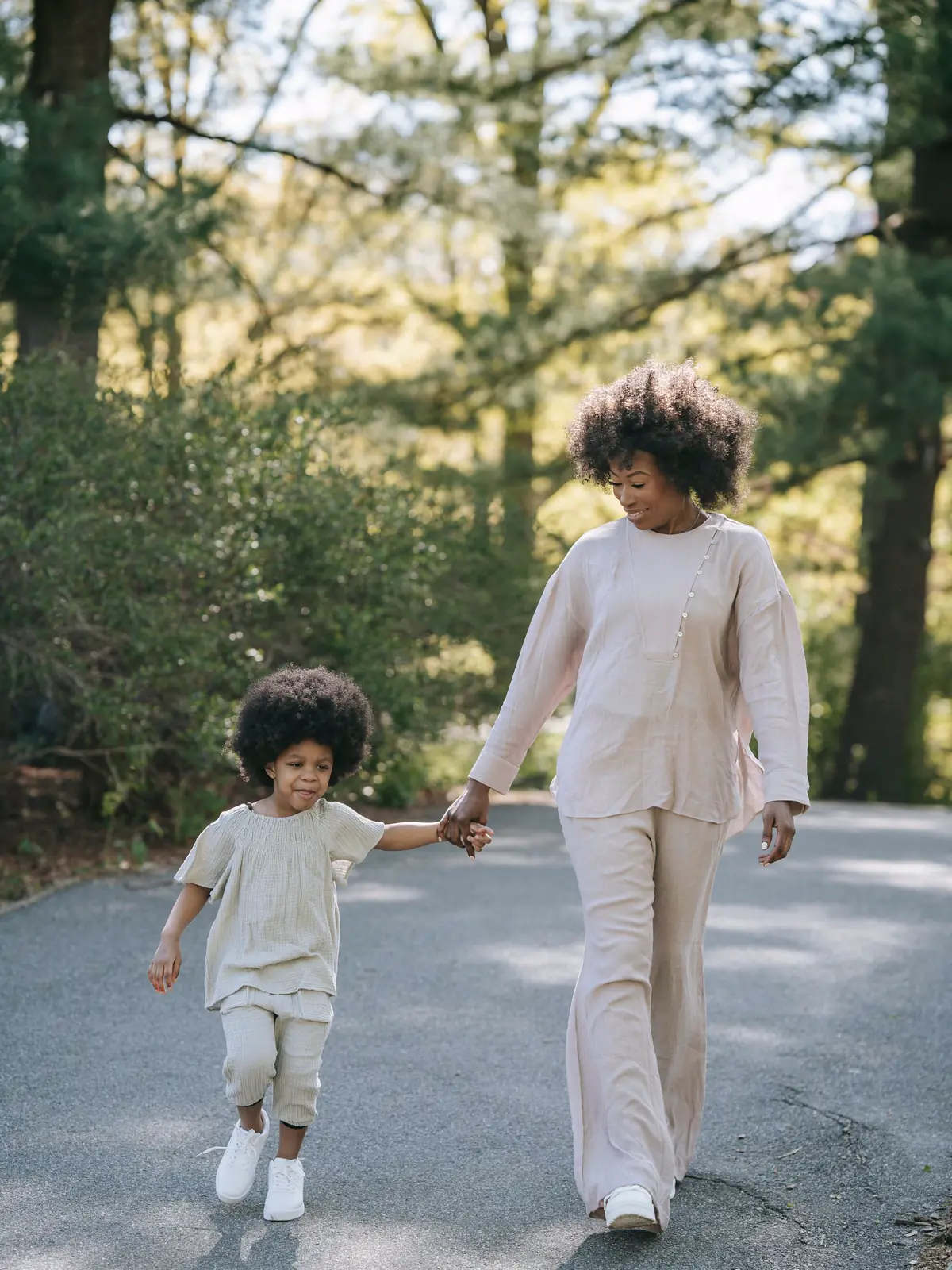 A mother and her young daughter walk hand in hand along a sunny park path