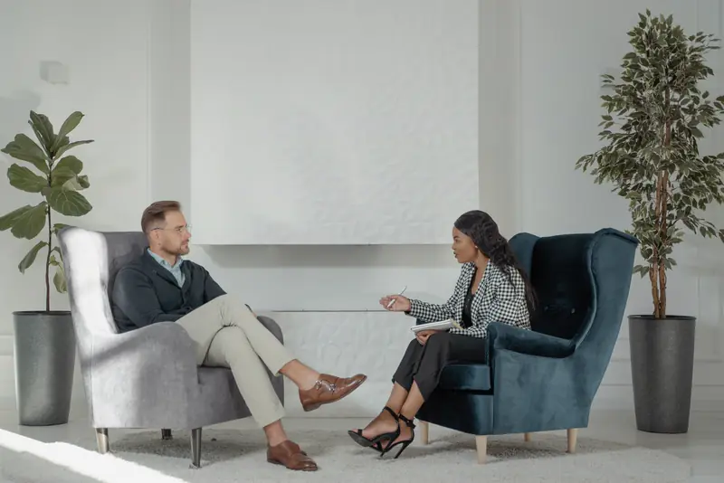 Two people talk in armchairs in a calm, plant-filled room