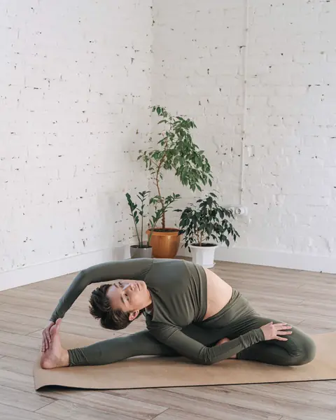 A woman stretches on a mat beside a tall window and houseplants.