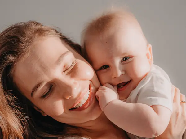 A smiling mother holds her baby cheek to cheek in warm sunlight.