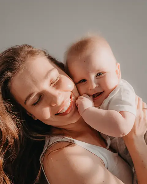 A smiling mother holds her baby cheek to cheek in warm sunlight.