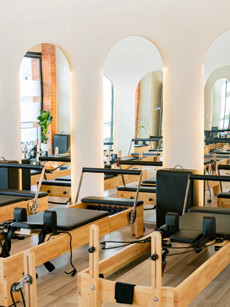 A bright studio with arched mirrors and a row of wooden Pilates reformers.