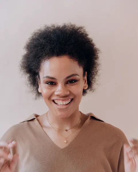 A woman with natural curly hair, in a tan sweater, laughs warmly.