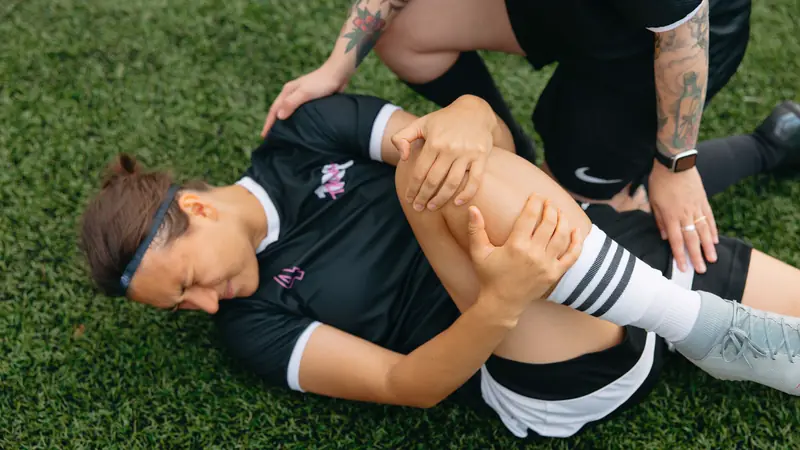 A soccer player lies on the grass holding her knee while a teammate helps.