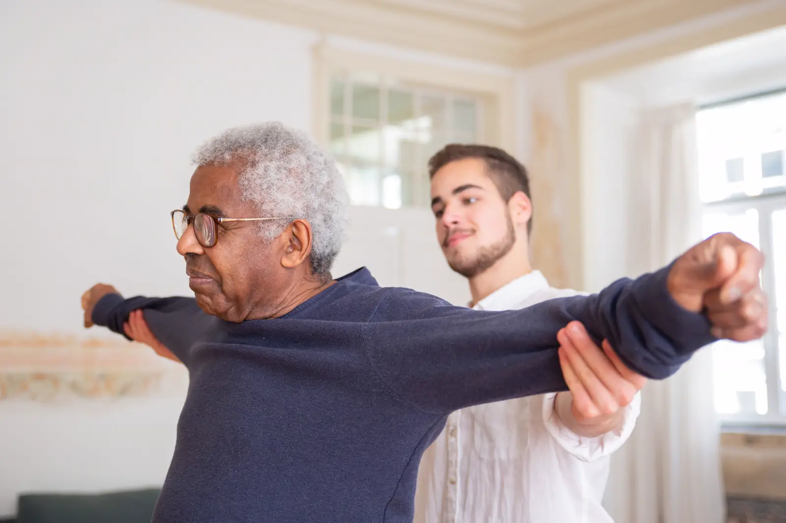 A therapist helps an older man stretch his arms out to the side.