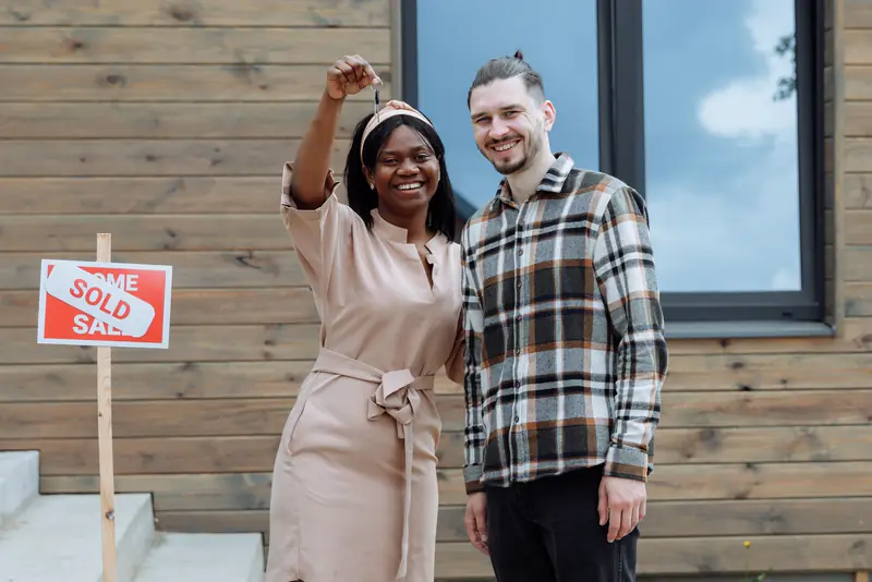 Two first-time buyers smiling and holding up their new keys beside a sold sign