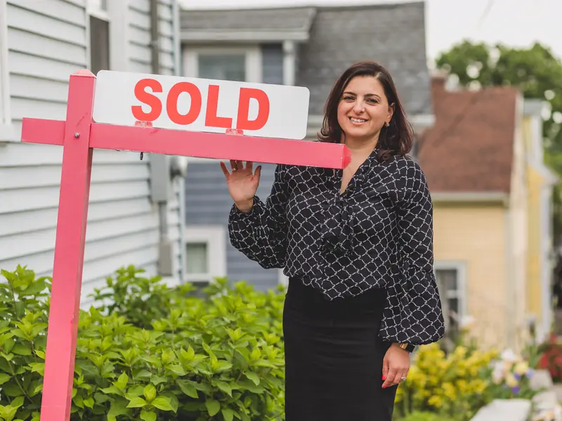 Dana Whitfield smiling beside a sold sign in front of a gray-sided house