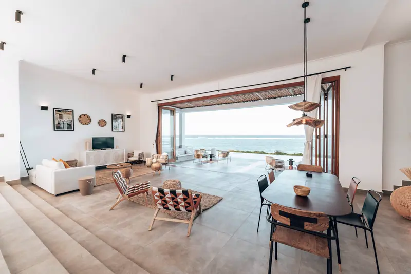 Dining table and woven chairs beside open glass doors and a view of the sea