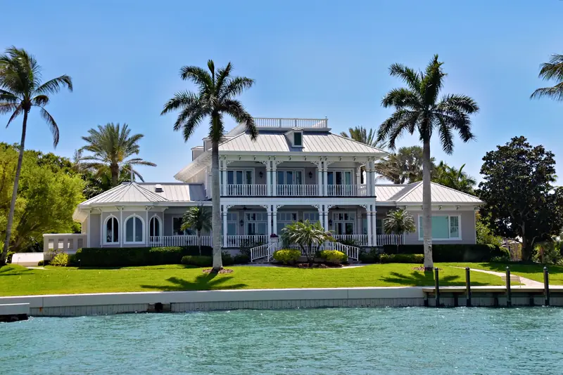 Two-story white home with deep porches, tall palms and a seawall on the water