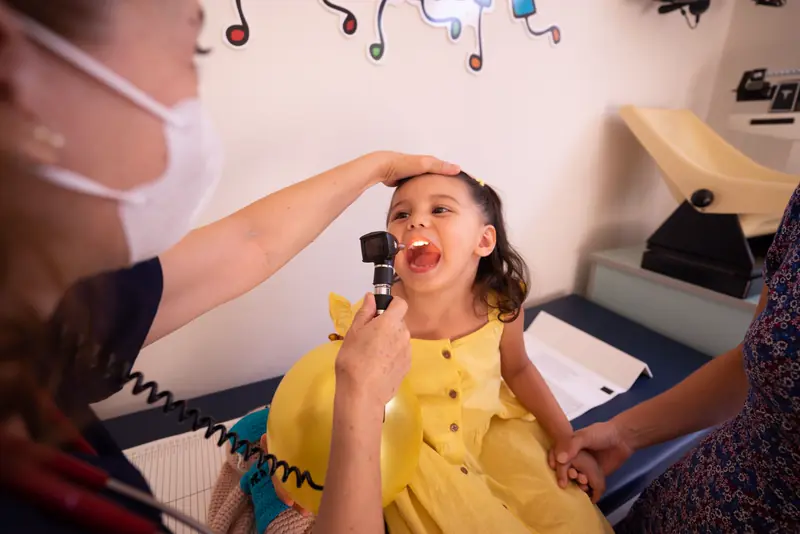 A smiling girl in a yellow dress says ahh for a throat check in a colorful exam room
