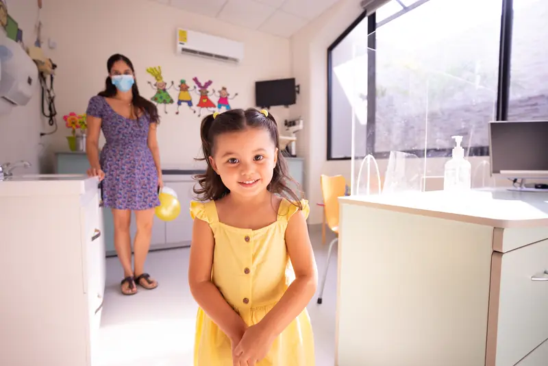 A smiling girl in a yellow dress stands in the colorful Little Lanterns exam room